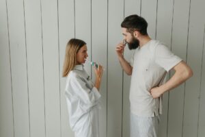 man and woman standing beside white wall while brushing their teeth