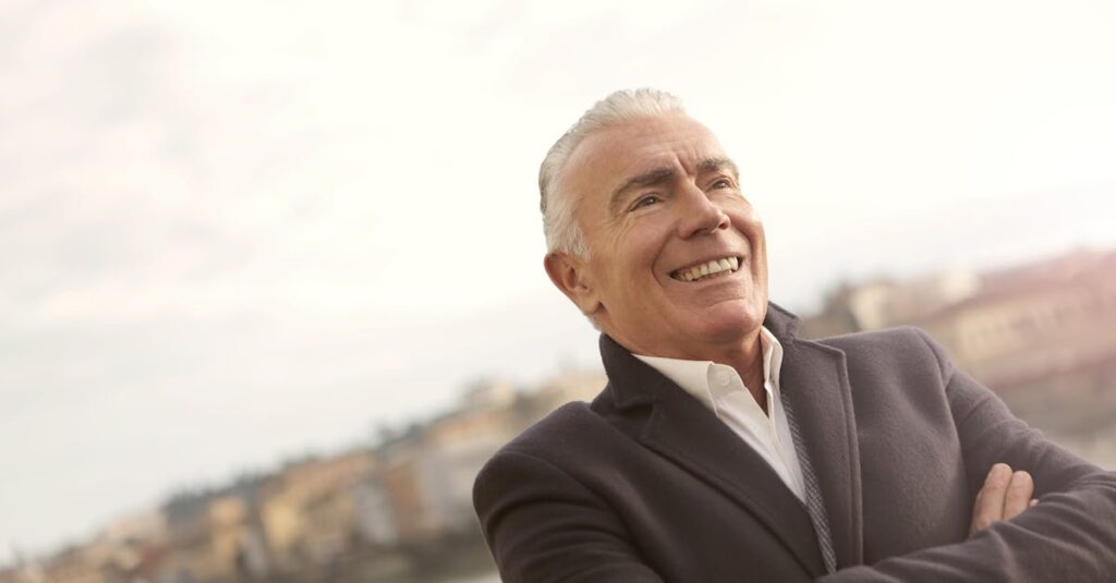 A senior man in business attire smiles confidently during a sunny outdoor setting.